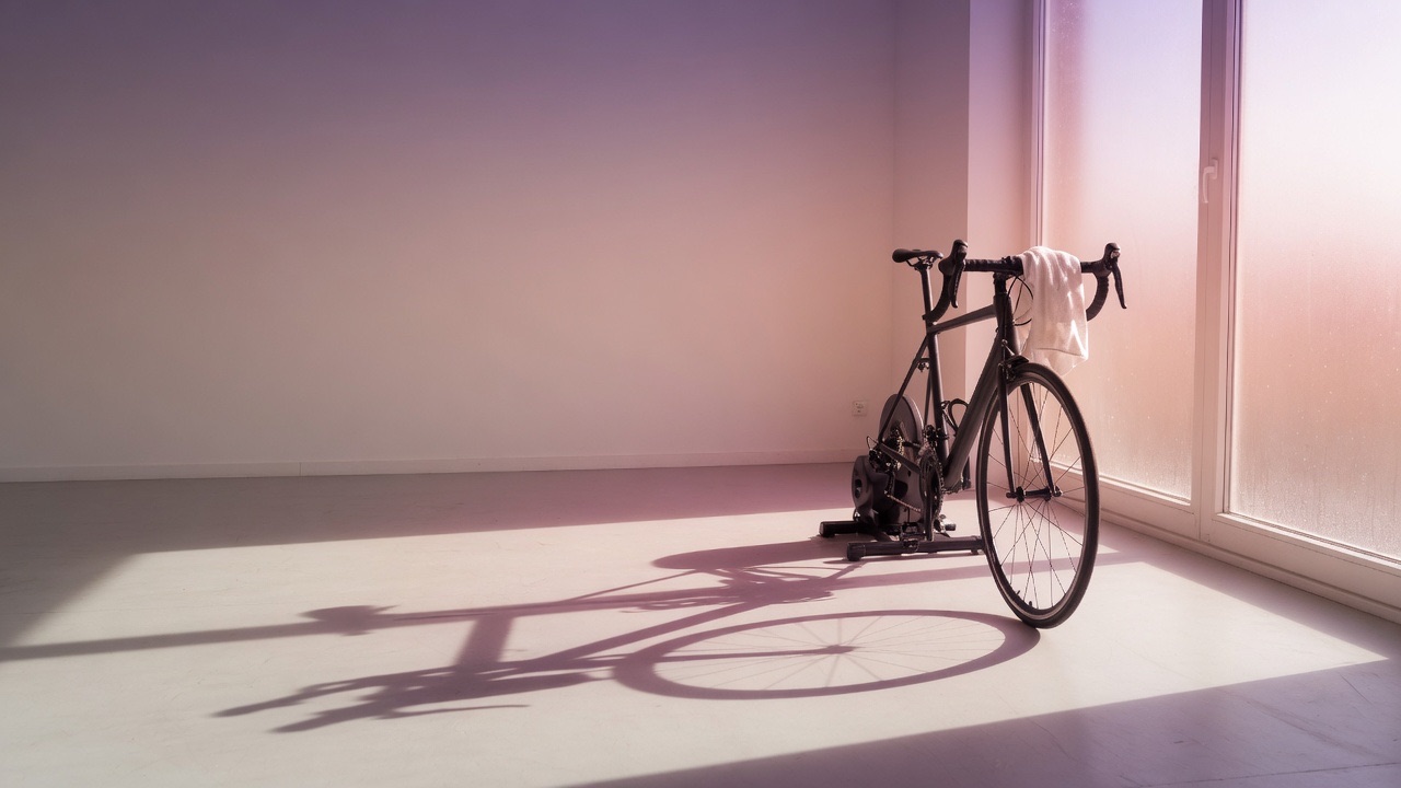 A bike on a trainer with a towel over the bars, morning light on the floor.